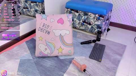 Media: Video of a pink, cloud-patterned cushion with a rainbow and unicorn design on a white and blue quilted floor, near a blue padded bench and a black keyboard.