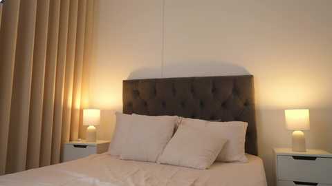 Media: Video of a modern bedroom with a tufted dark gray headboard, white bedding, two white pillows, beige curtains, and matching nightstands with warm-toned lamps.