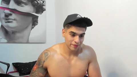 Media: Video of a shirtless, muscular, tan-skinned man with a black cap, wearing a black and red shirt, tattoos on his left arm, in a minimalist room with a black and white photo of a classical statue on the wall.