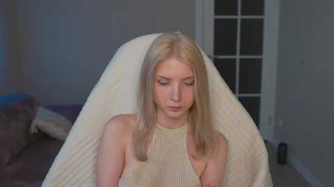 Media: A video of a young, fair-skinned, blonde woman with long hair wearing a beige, sleeveless top, sitting on a white sheet in a dimly lit bedroom with gray walls and a partially open door.