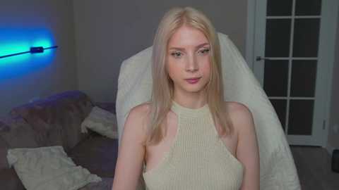 Media: Video of a young woman with light skin and long, straight blonde hair, wearing a cream-colored, ribbed sleeveless top, sitting on a white fur blanket in a dimly lit room with a blue LED strip on the left and a glass door on the right.