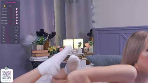 Media: Video of a woman in a white top, lying on a bed, wearing white socks, with a VR headset on, looking at a virtual room with a mirror, flowers, and books.