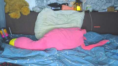 Media: Video of a pink flamingo plush toy sleeping on a blue bedspread, surrounded by stuffed animals and a lantern in a cozy bedroom.