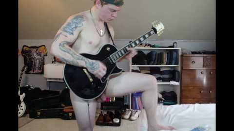 Media: Video of a muscular, inked man with fair skin, wearing only a green cap, playing a black electric guitar in a cluttered bedroom.