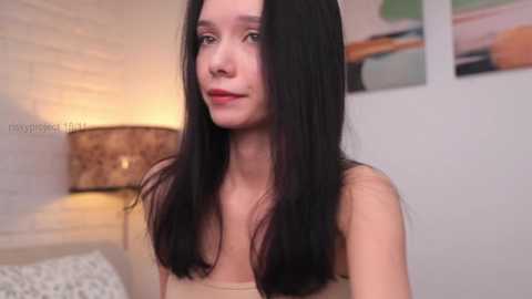 Media: Video of a young woman with long, straight black hair, fair skin, and a slender physique, wearing a beige tank top. She's indoors with a modern, white-walled room in the background, featuring abstract art and a beige lampshade.