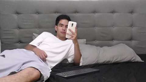 Media: A video of a young Asian man with short black hair, wearing a white T-shirt and grey sweatpants, lying on a grey tufted headboard bed, taking a selfie with a white iPhone.