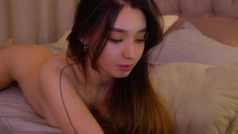 Media: Video of a nude young woman with long, dark hair lying on a bed, wearing black earbuds, her face turned slightly to the side, in a softly lit bedroom.