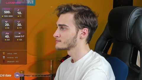 Media: A video of a young man with short, light brown hair and a beard, wearing a white t-shirt, seated in a black gaming chair, watching a streaming video featuring statistics on the left screen.