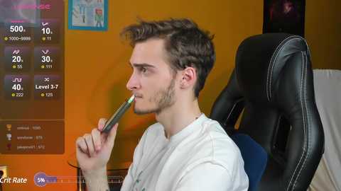 Media: Video of a young man with short, wavy brown hair, wearing a white t-shirt, sitting in a black gaming chair. He holds a green pen to his mouth, looking thoughtful. Background shows a yellow wall with a framed map, and a virtual chat window on the left.