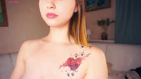 Media: Video of a topless young woman with light skin, light brown hair, pink lips, and a tattoo of a red heart with paw prints on her shoulder, set in a cozy, softly lit room with a white sofa and blue curtains.