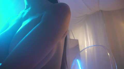 Media: A video of a woman's bare back, with a glass of wine in the foreground, under soft blue and yellow lighting.