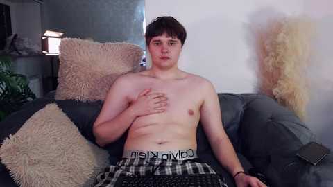 Media: Video of a shirtless, pale-skinned, chubby young man with short brown hair, wearing Calvin Klein boxers, sitting on a gray couch in a dimly lit room.