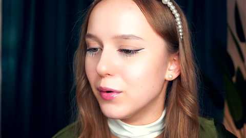 Media: Video of a young woman with light skin, long straight brown hair, and a pearl headband. She wears a white high-collared top and has subtle makeup with winged eyeliner. Dark curtains form the background.