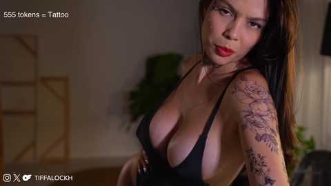 Media: Video of a woman with long, dark hair, wearing a low-cut black dress, revealing ample cleavage. She has tattoos on her arm and a confident expression. Background shows dimly lit room with a green plant.