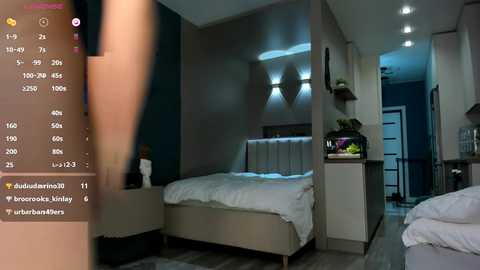 Media: Video of a modern, minimalist bedroom with beige walls, a white bed, a green plant, and a sleek nightstand. A blurred person's leg is partially visible on the left.