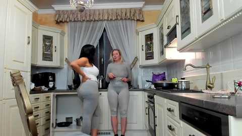 Media: Video of a modern kitchen with two women in matching grey workout outfits, one facing the other. White cabinets, beige walls, and a window with sheer curtains.