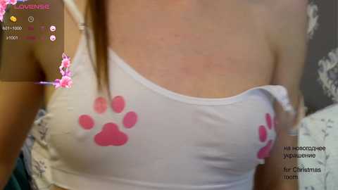 Media: Video of a woman in a white tank top with pink paw prints, revealing her small breasts through the thin fabric. The background includes a blurry room with floral patterns and festive Christmas decorations.