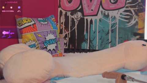 Media: Video of a plush, anthropomorphic penis-shaped toy with exaggerated, large testicles lying on a bed, surrounded by colorful comic-style pillows and graffiti-covered wall, creating a playful and slightly risqu\u00e9 atmosphere.