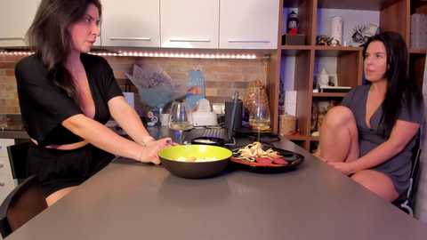 Media: Video of two women in a modern kitchen; one preparing pasta, the other seated at the counter in casual attire, both with medium build, dark hair, and light skin.