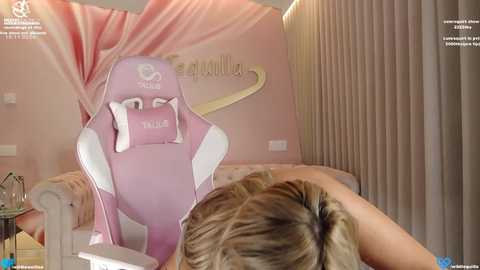 Media: Video of a person with blonde hair, wearing a pink and white gaming chair, in a cozy, pastel-pink bedroom with curtains and soft lighting.