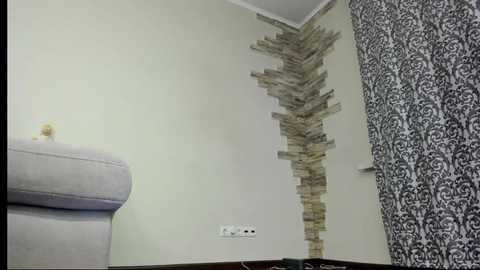 Media: A video of a modern, beige-walled room with a dark wood floor. On the left, a light grey armchair. On the right, a wall with a vertical arrangement of stacked, textured stone tiles. A black and white patterned curtain hangs in the background.