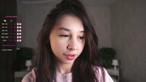 Media: Video of an East Asian woman with long, straight black hair and fair skin, wearing a light pink top. Background features a white wall with potted plants and a TV screen displaying streaming stats.