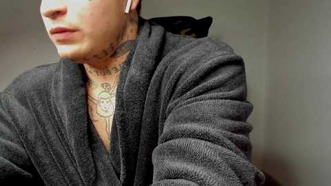 Media: Video of a person with pale skin, wearing a gray plush robe, white earbuds, and visible tattoos including a heart and script on the chest.