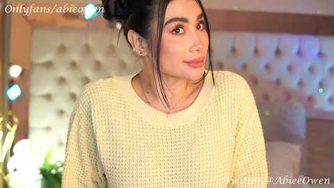 Media: Video of a young woman with dark hair in a bun, wearing a cream-colored knitted sweater, in a modern room with a plush, tufted headboard and soft lighting.