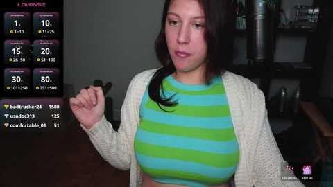 Media: Video of a young woman with long dark hair, wearing a green and blue striped crop top and a white cardigan, holding a microphone, in a dimly lit room with a black background.