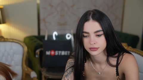 Media: Video of a young woman with long black hair, wearing a black top, sitting on a green velvet couch. Background includes a lamp, floral wallpaper, and a black \"HOTTEST\" shopping bag.