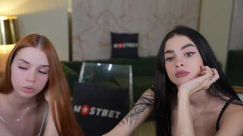 Media: Video of two young women with long hair, one red-haired and one black-haired, sitting on a green couch. Background includes a black \"MOST BET\" sign, a beige wall, and a lamp.