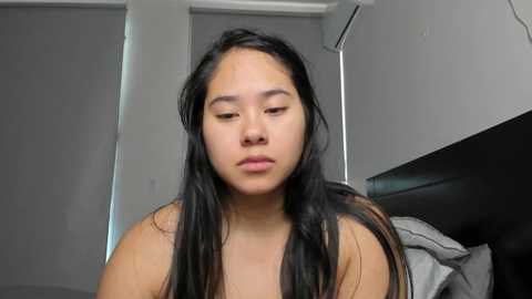 Media: Video of an Asian woman with long black hair, light skin, and closed eyes, sitting indoors with a grey bed and white walls in the background.