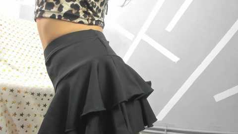 Media: Video of a woman from the waist down, wearing a leopard print crop top and a black pleated skirt, standing in a modern room with a white ceiling and a patterned chair.