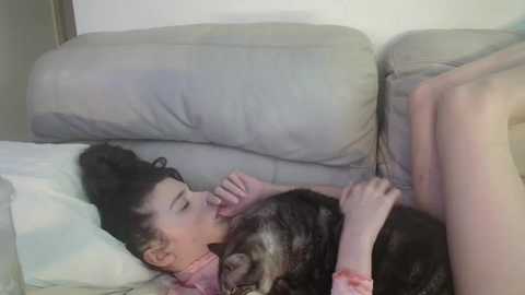 Media: Video of a young Asian girl with dark hair in a ponytail lying on a beige sofa, kissing a tabby cat. She wears a pink shirt.