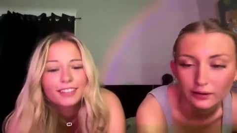 Media: Video of two smiling young women with light skin, one with blonde hair, the other with brown hair, wearing sleeveless tops, seated indoors against a dark curtain background.