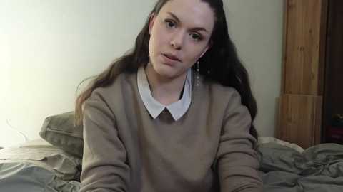 Media: Video of a young woman with long brown hair, wearing a beige sweater and white collared shirt, sitting on a bed with gray sheets in a dimly lit room.