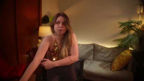 Media: A video of a slender, fair-skinned woman with long brown hair, wearing a gold necklace, seated on a black chair in a dimly lit living room with a beige couch, wooden furniture, and a potted plant.