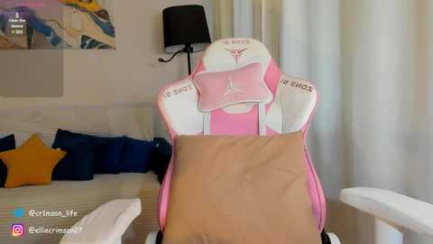 Media: A video of a pink and white gaming chair with a beige cushion, positioned in a cozy living room with blue and yellow pillows on a couch, a black lamp, and a colorful abstract painting on the wall.