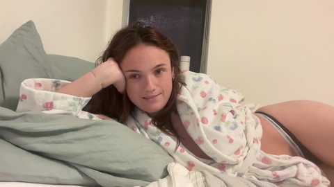 Media: Video of a smiling young woman with fair skin, long brown hair, and a medium build, lying on a bed in pajamas, surrounded by green and white bedding.