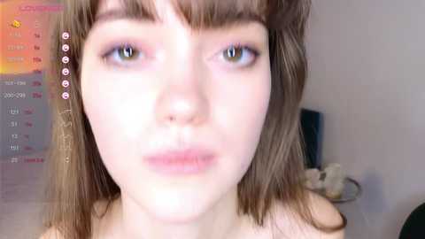 Media: Video of a young woman with fair skin, light brown hair, and brown eyes. She has a neutral expression, wearing minimal makeup. Background includes a calendar and a blurred wall.