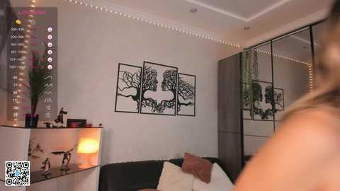 Media: Video of a modern bedroom with three minimalist, black-and-white tree illustrations on a white wall. A black couch with beige pillows is visible, accompanied by a small, lit-up lamp and a QR code in the lower left corner.