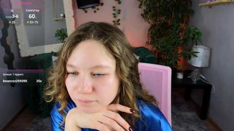 Media: Video of a young Caucasian woman with curly brown hair, wearing a blue shirt, sitting in a pink chair, looking introspective. Background features a green couch, potted plant, and gray walls.