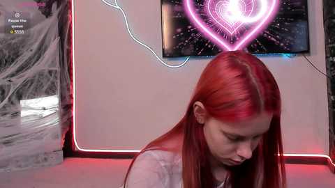 Media: A video of a young woman with straight red hair, wearing a white top, sitting in front of a wall with a heart-shaped neon sign and a digital clock.