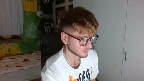 Media: Video of a young man with short, spiky, dyed hair and glasses, wearing a white t-shirt, sitting on a chair in a cluttered room with a football helmet and inflatable.