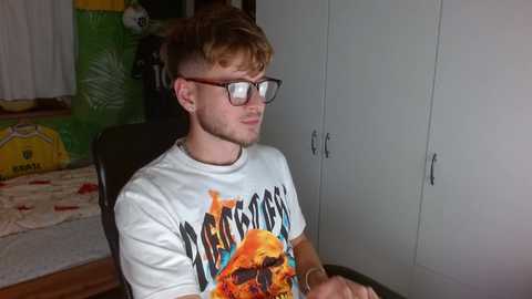 Media: Video of a young man with short, tousled brown hair, wearing black-rimmed glasses and a white \"Mars Attacks\" t-shirt, sitting on a dark chair, with a green inflatable toy in the background.