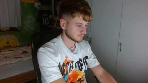 Media: Video of a young man with short, light brown hair, wearing a white T-shirt featuring an orange and black graphic design, sitting on a dark chair in a cluttered, dimly lit room with a green wall and a yellow and white rug.