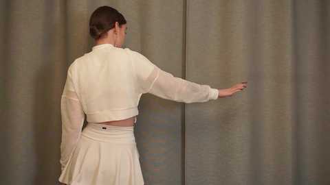 Media: Video of a woman with light skin and dark hair in a bun, wearing a cream-colored, semi-transparent, long-sleeved crop top and pleated skirt, standing against a muted green curtain, reaching out with her right hand.