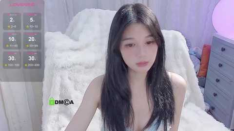 Media: Video of an East Asian woman with long black hair, light skin, wearing a light blue tank top, sitting on a white fur rug in a softly lit room with a gray dresser and orange pillow in the background.