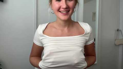 Media: Video of a smiling, fair-skinned woman with light brown hair tied back, wearing a tight white T-shirt that reveals her small breasts. She stands in a white-tiled bathroom with a towel rack in the background.