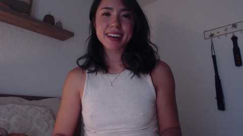 Media: Video of a smiling woman with medium-length black hair, wearing a white tank top, standing in a dimly lit bedroom with a wooden shelf and hanging black items on the wall.